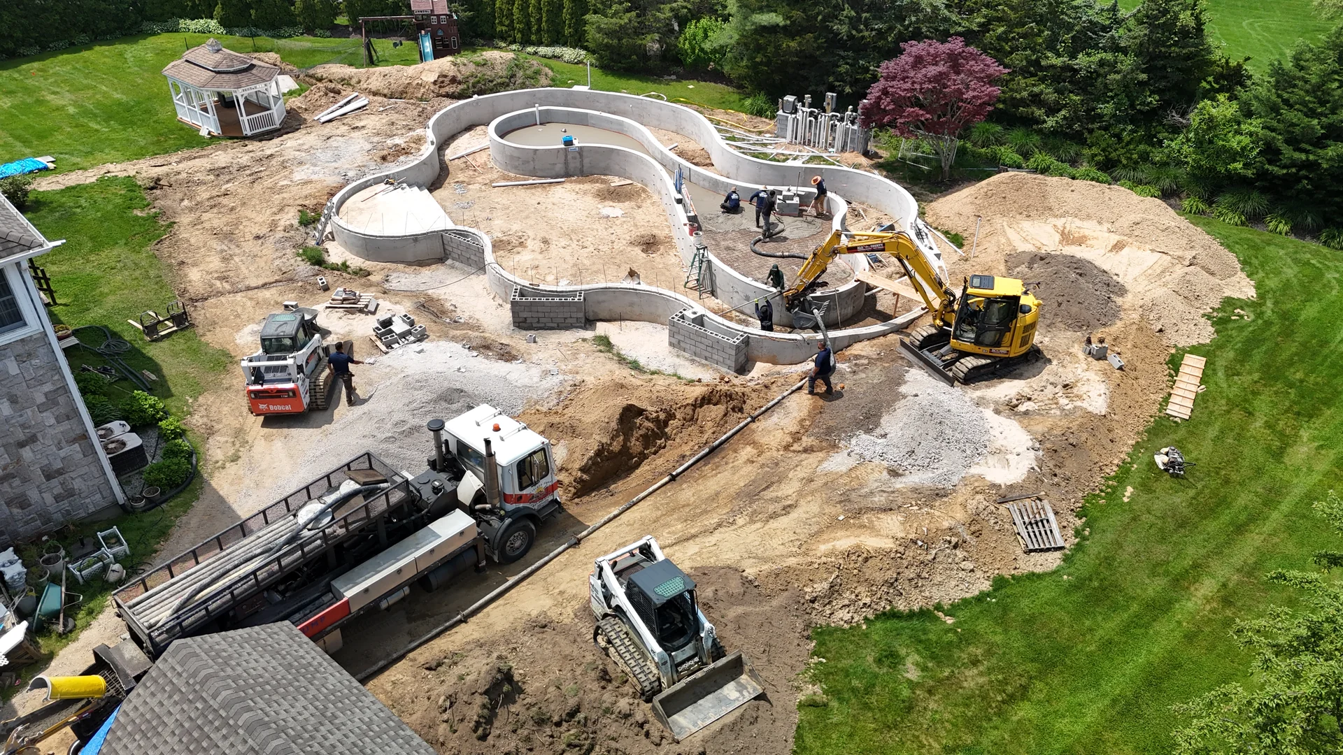 Aerial of the full lazy river shell formed in open ground, with excavator, skid steers, concrete truck and crew working the site, East Northport