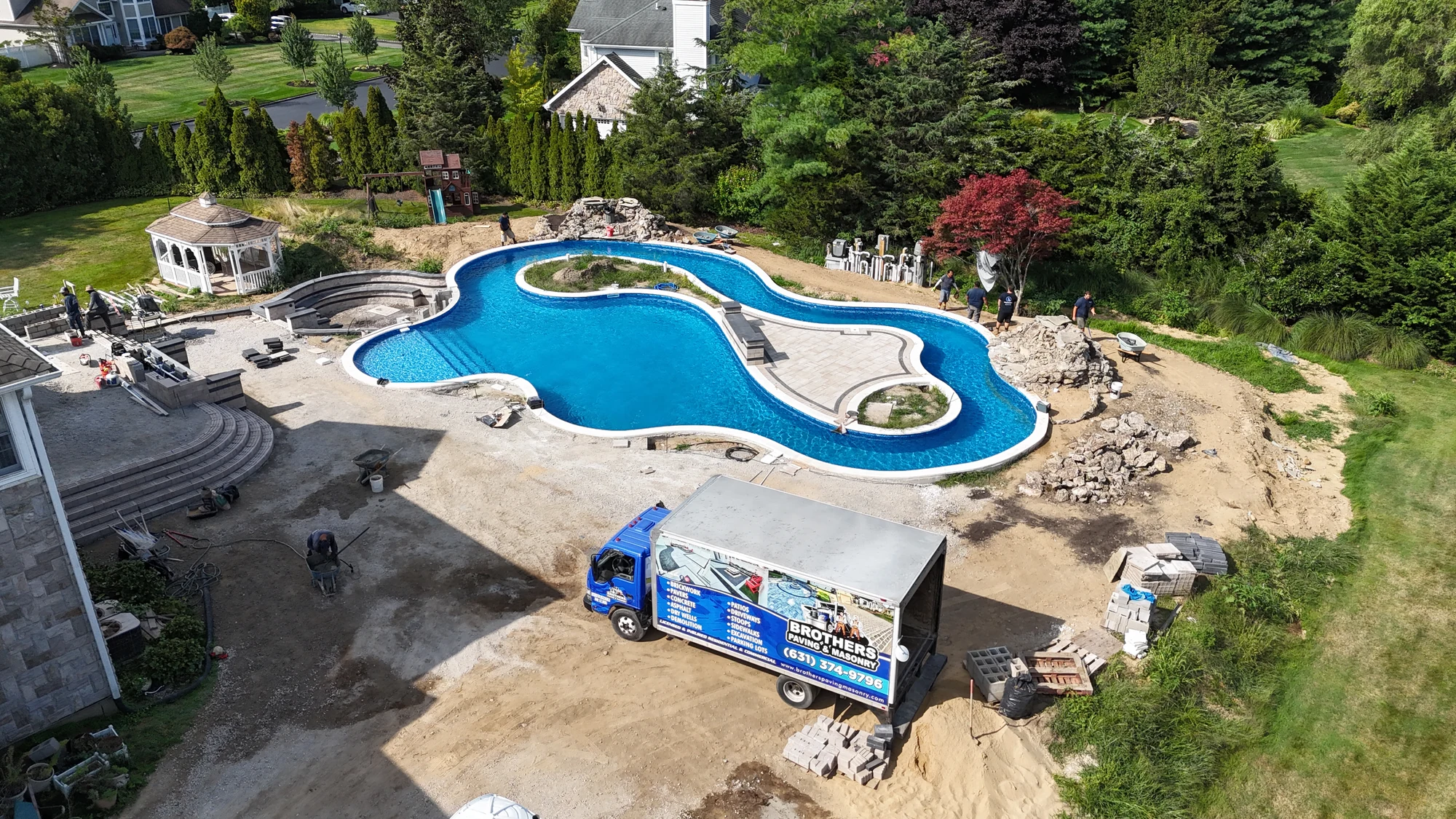 Brothers Paving & Masonry box truck parked beside the lazy river mid-build, with crews working around the pool, East Northport
