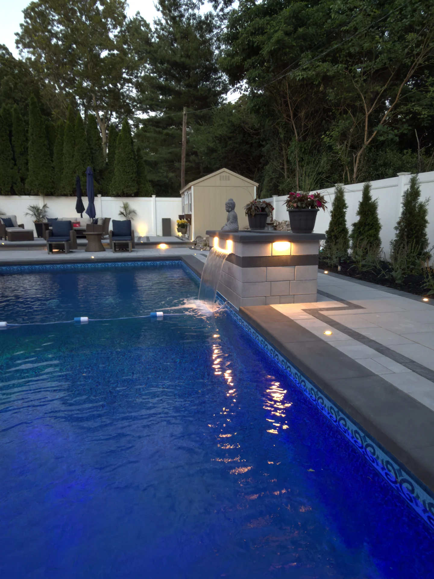The raised spa spilling into the lit pool over a sheer descent at dusk, Nesconset