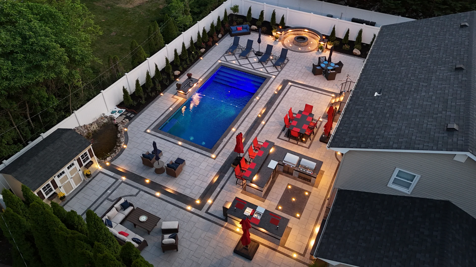 Blue-hour aerial of the lit pool, raised spa, outdoor kitchen and fire terrace in Nesconset, Long Island